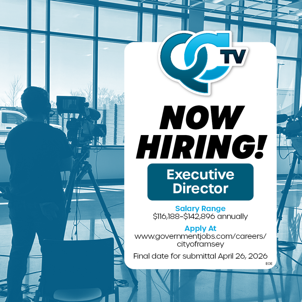 A room with large glass windows, on the left, a man standing behind a professional view camera. On the right, white box at the top, QCTV logo, words below, Now Hiring! Executive Director, Salary Range $116,188–$142,896 annually, Apply at www.governmentjobs.com/careers/cityoframsey Final date for submittal April 26, 2026